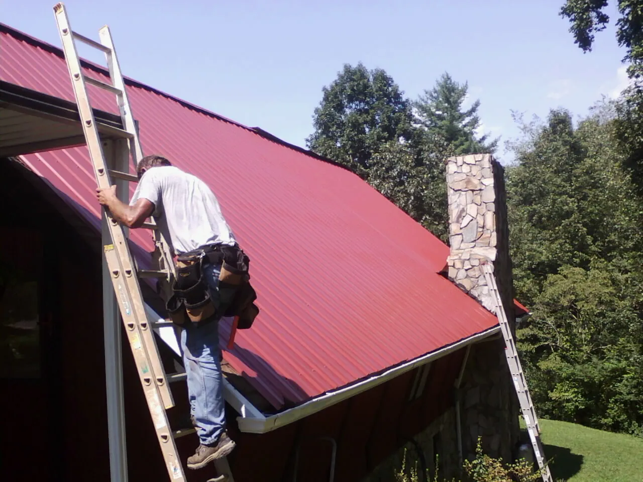 Roofer on a ladder beside a red metal roof and stone chimney — Soffit & Fascia Repair by Roofing Brothers in Colwell